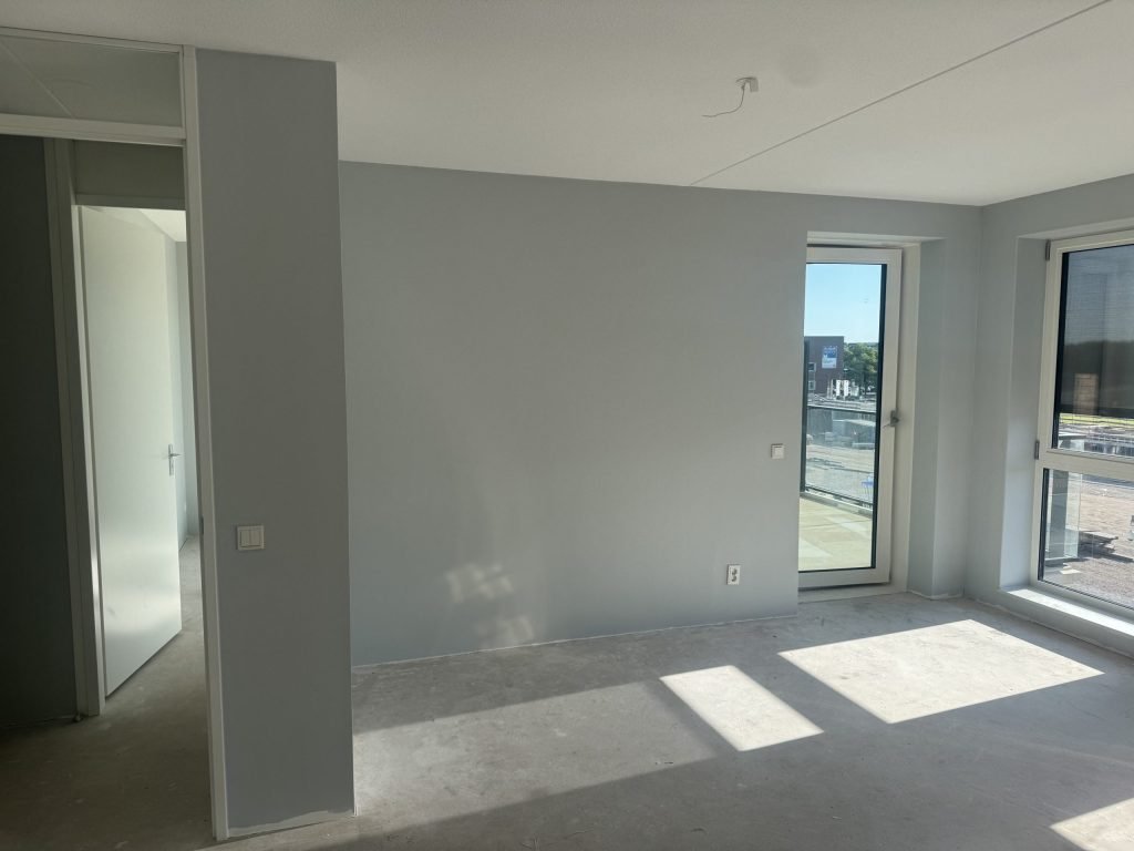 Net opgeleverd appartement met lichtgrijs geschilderde wanden, balkondeur en uitzicht op een bouwterrein met nieuwe woonblokken
